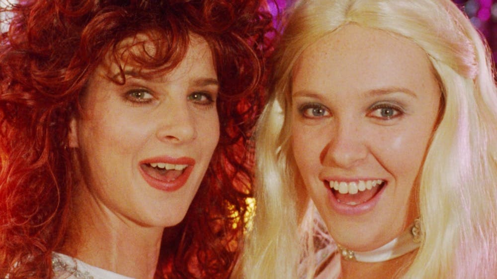 Two women look directly at the camera midway through song, woman on left with big curly hair