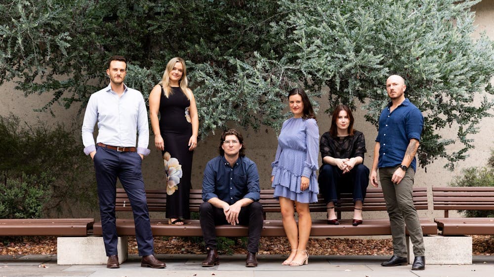 Five adults sitting on or standing around a bench outside.