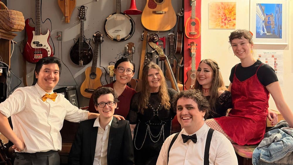 7 dapperly-dressed performers (4 women, 3 men) posing in front of a wall of musical instruments