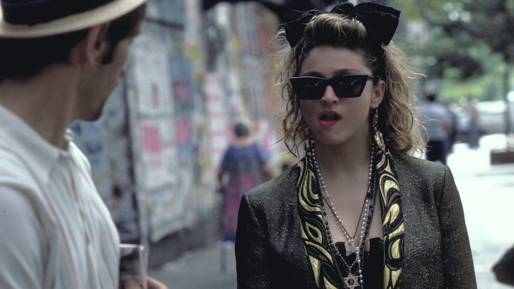 Woman walking towards man wearing 1980s style clothing, sunnies and big hair bow