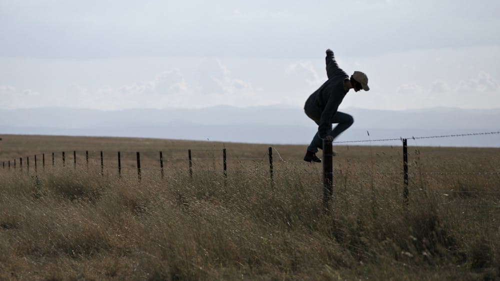 A man in a flat field jumping up onto a timber pole for fencing