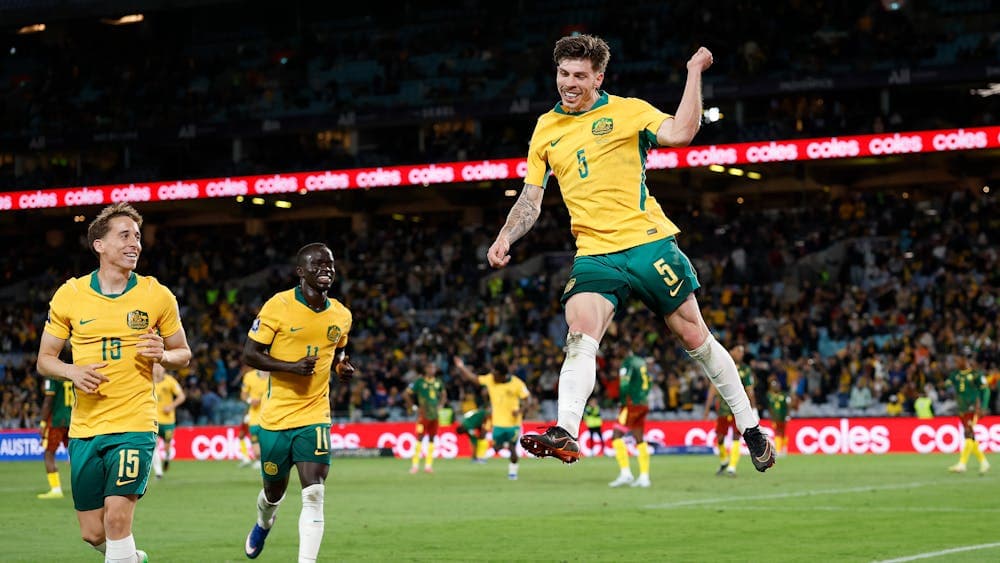 Soccer players in yellow kits celebrate a goal on a stadium pitch as teammates run in behind