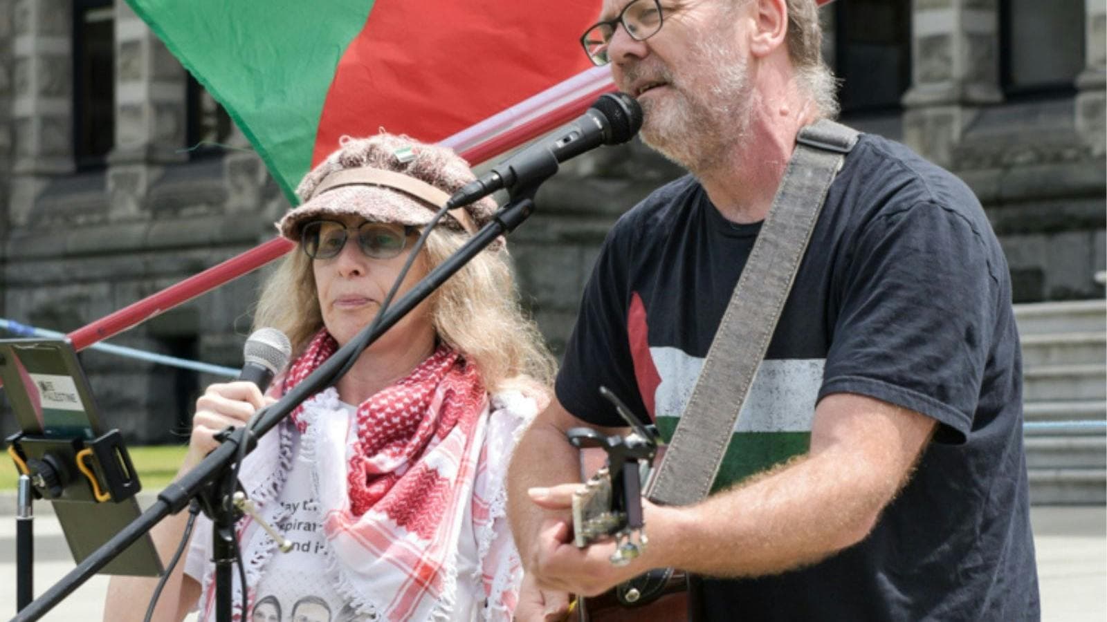 David Rovis & Kamala Emanuel singing live at a protest in front of a Palestinian flag.]