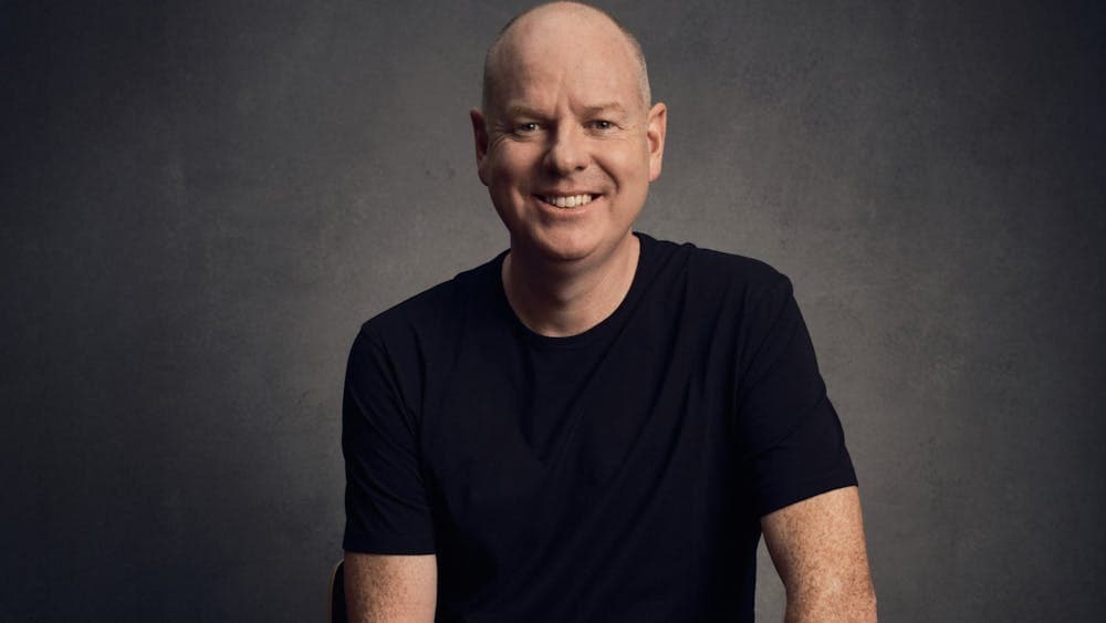 Comedian Tom Gleeson sitting on a stool, smiling