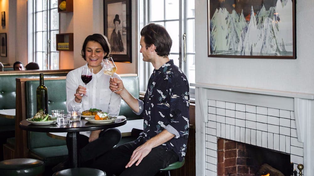 Couple with food and wine on tall stools by a fire place