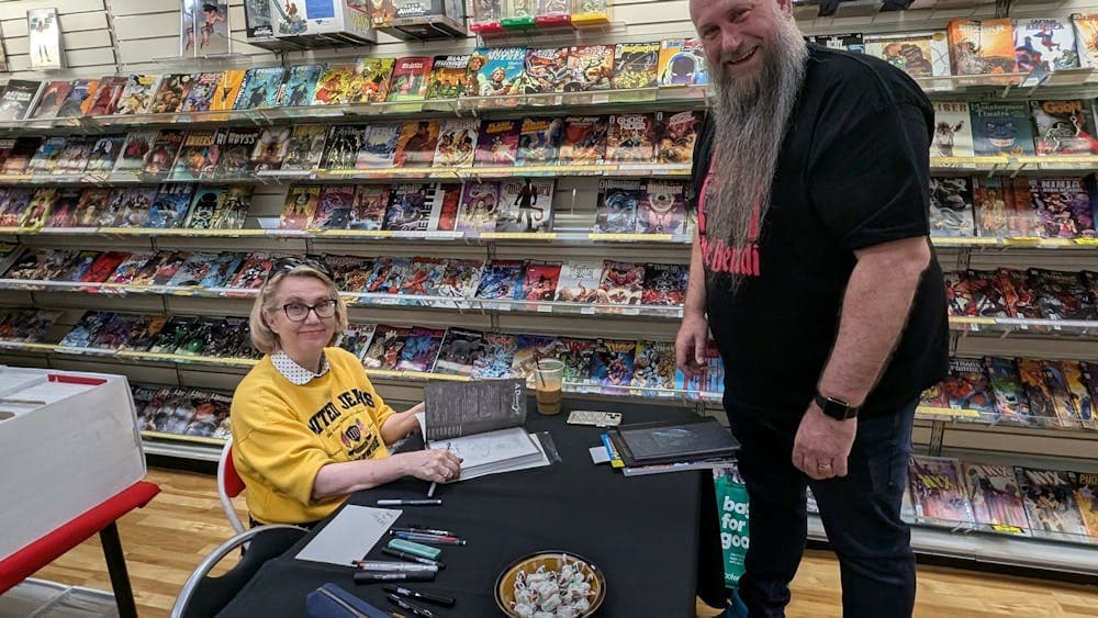 A woman sits at a table with signed books and pens, a man standing waits. Both smiling at the camera