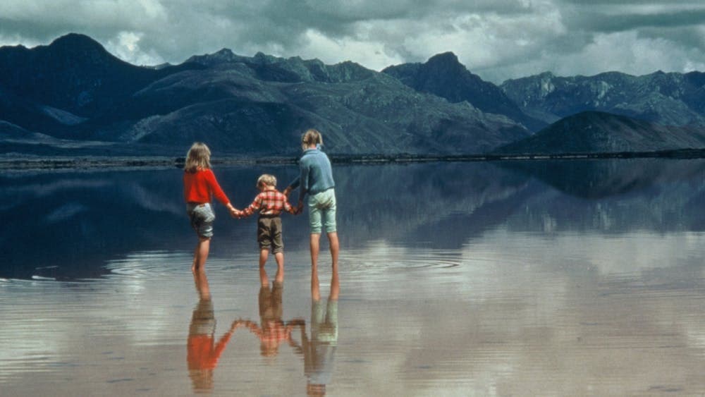 Three children holding hands standing in water with mountains in background