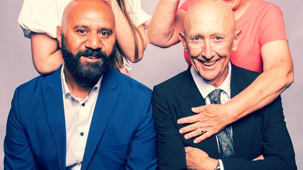 Two men sitting and theor wives standing behind them making silly faces