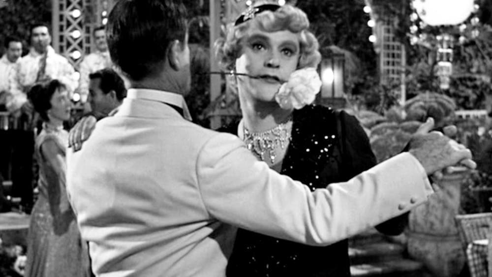 Black and white still of two men dancing, one dressed as a woman with a flower in his mouth