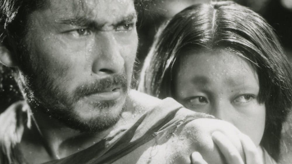 Black and white still of a man looking angry and a woman hiding behind the man