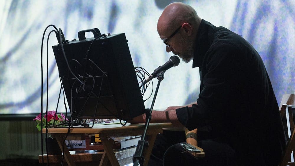 Person performs live electronic music with microphone and equipment before a large abstract projecti
