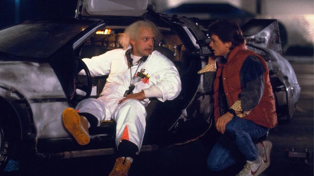 Two men facing each other, man on left sitting in car in white doctors suit
