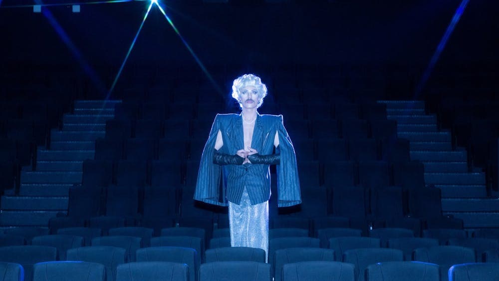 Drag artist stands in the middle of an empty cinema wearing an overcoat and blonde wig