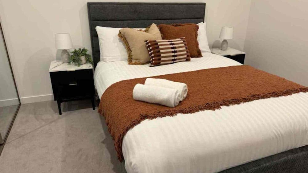 Bedroom with grey fabric headboard, white linens, rust-orange throw blanket, and black bedside table