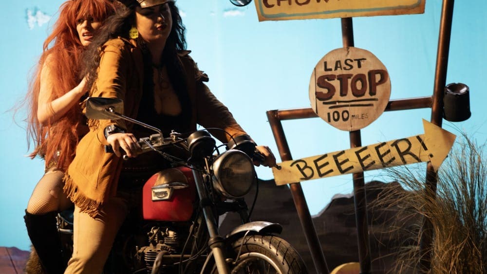 Two women dressed in costumes and wigs on a motorcycle in a theatre like setting