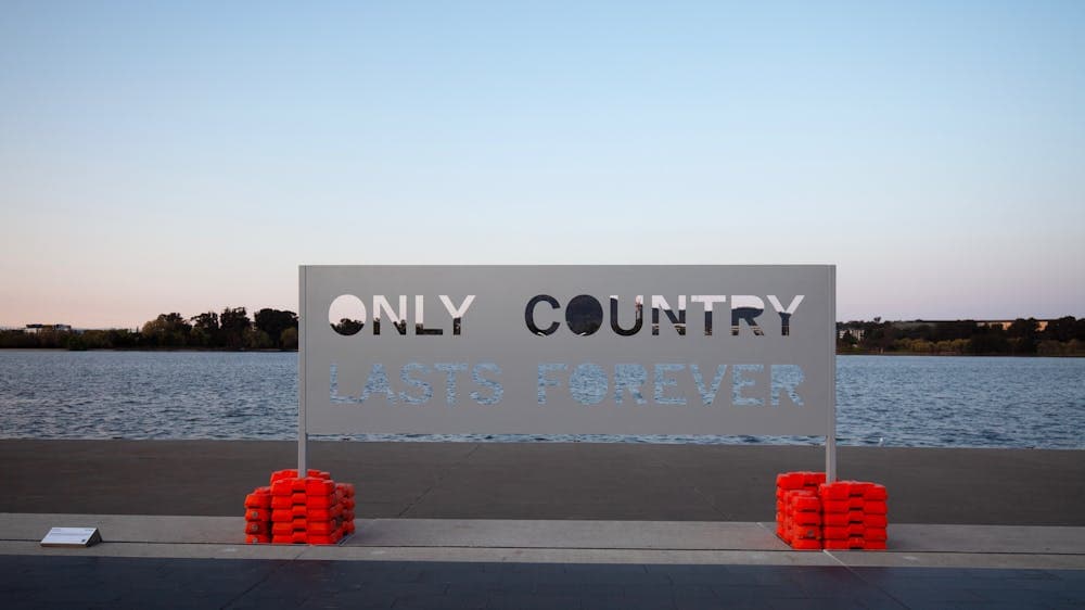 A metal sign that reads "Only Country Lasts Forever" positioned in front of Lake Burley Griffin.