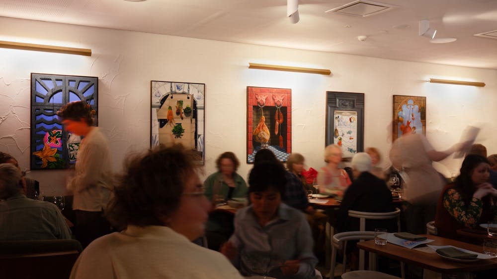 A restaurant full of people moving, with colourful paintings hung on the walls.