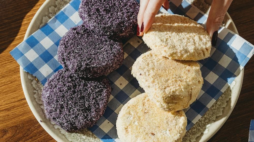A plate of purple and beige pastries on checkered paper, placed on a wooden table with two cups of c