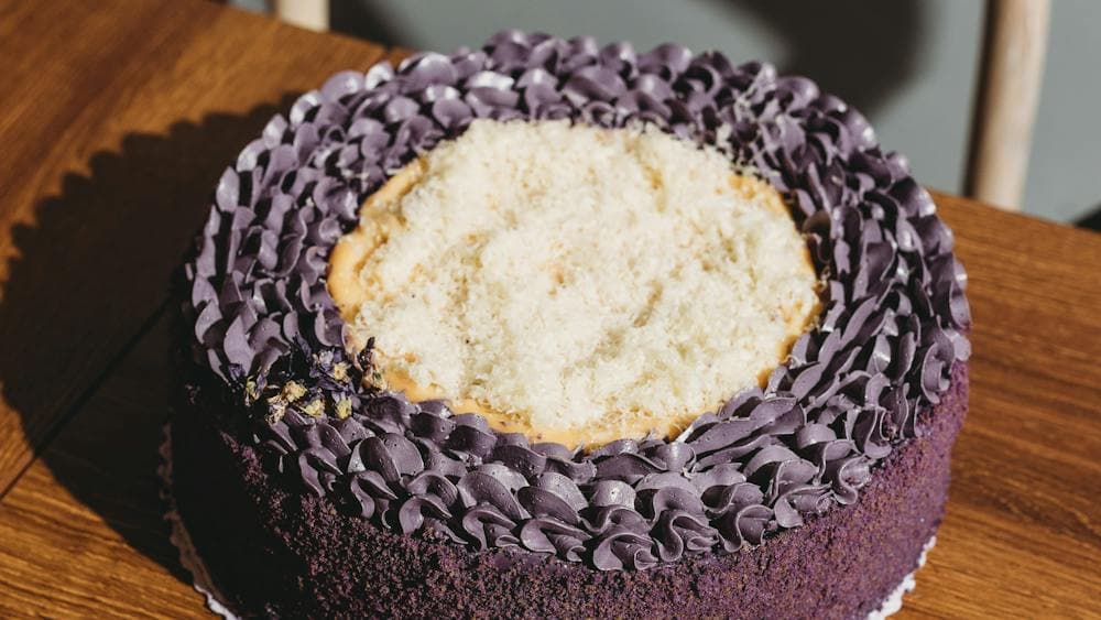 A round ube cake with purple frosting and a light shredded topping, placed on a wooden surface.