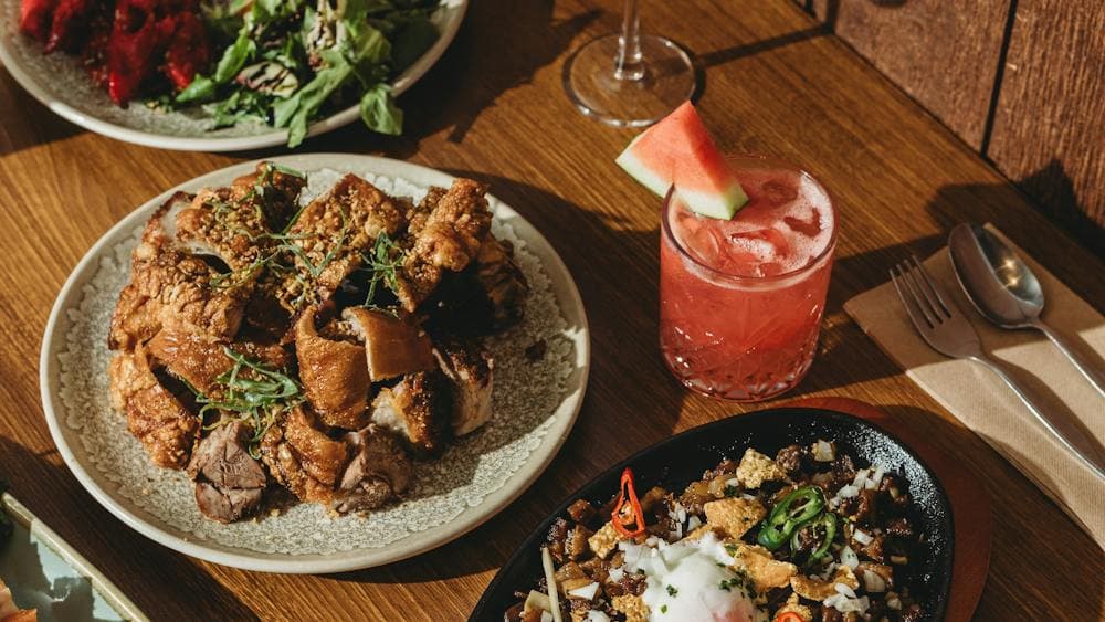 A wooden table filled with Filipino dishes including sizzling sisig with egg, crispy pork pieces, a