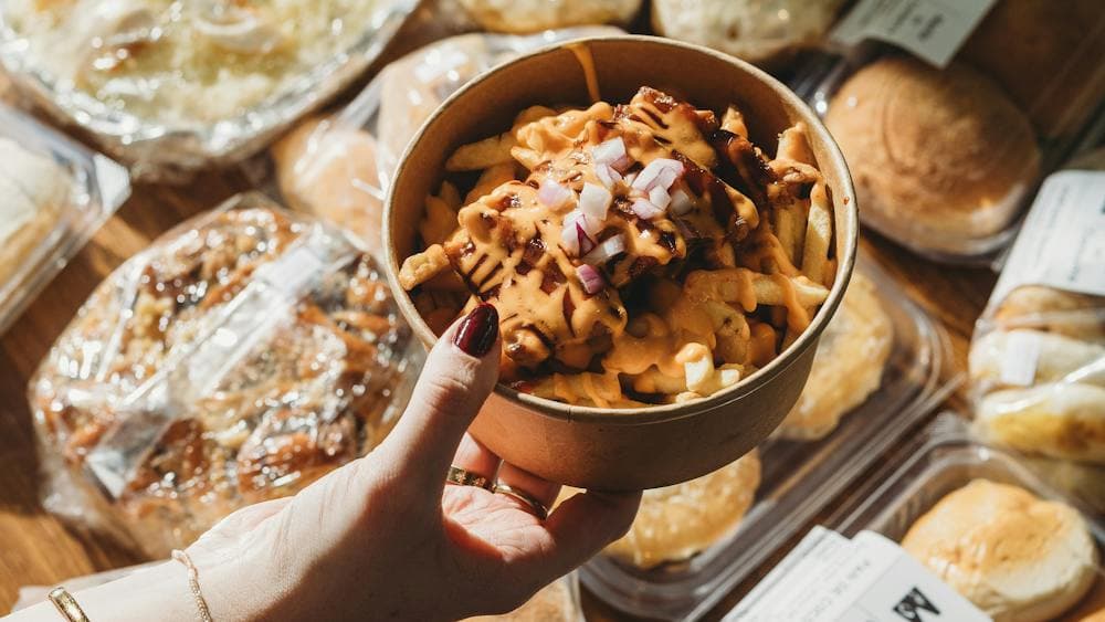 A hand holding a bowl of lechon loaded fries topped with cheese and onions, surrounded by assorted F