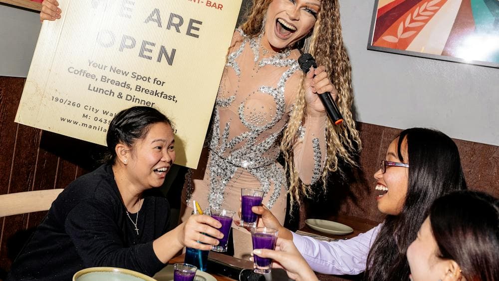 A performer holding a microphone stands beside a table where guests raise purple drinks in a toast,