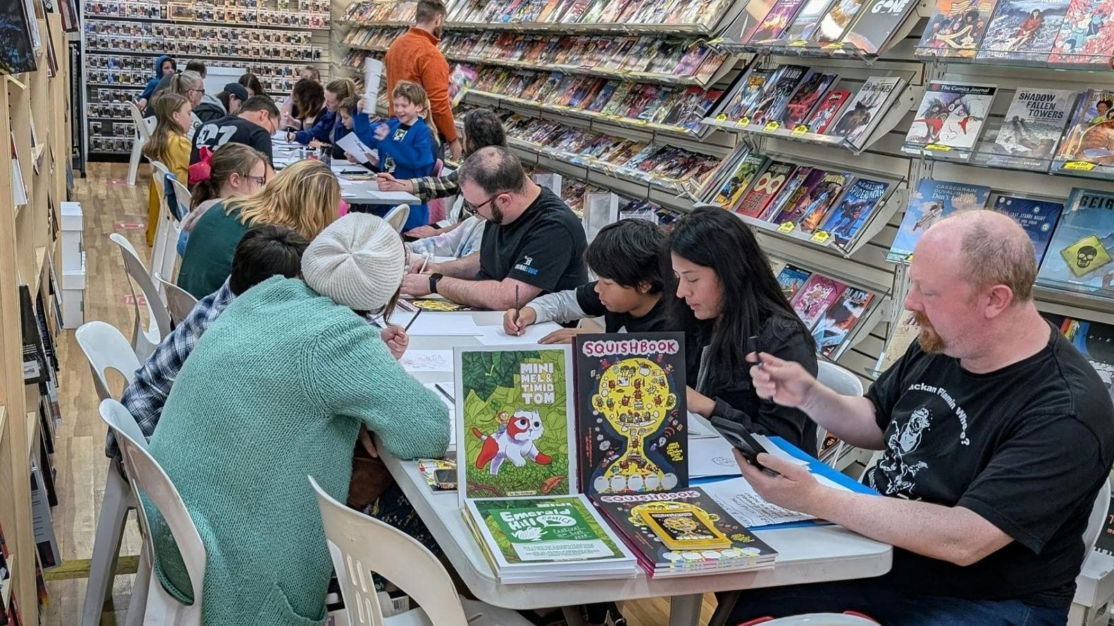 Adults and children sitting at tables in a comic shop drawing on paper
