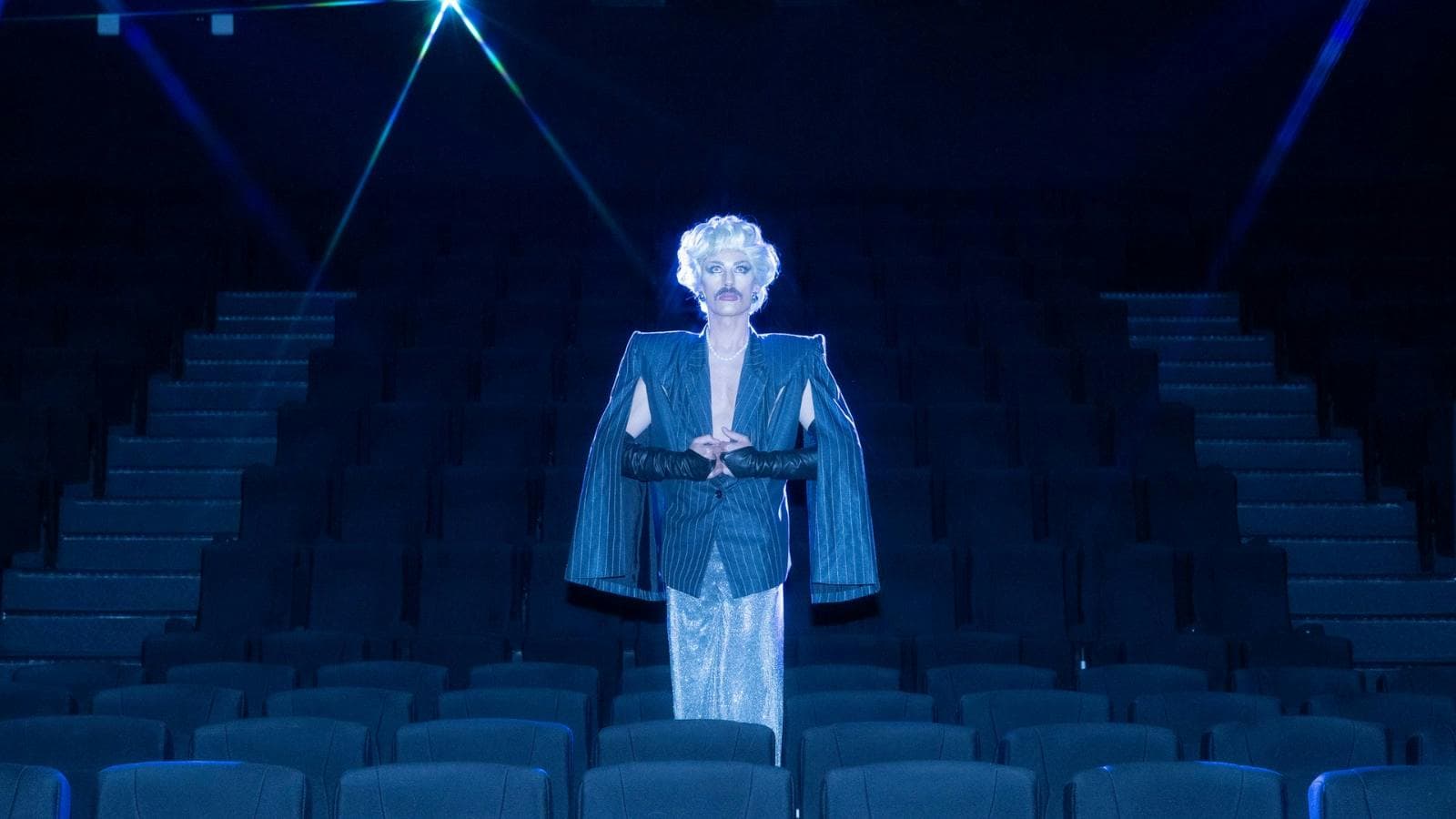 Drag artist stands in the middle of an empty cinema wearing an overcoat and blonde wig