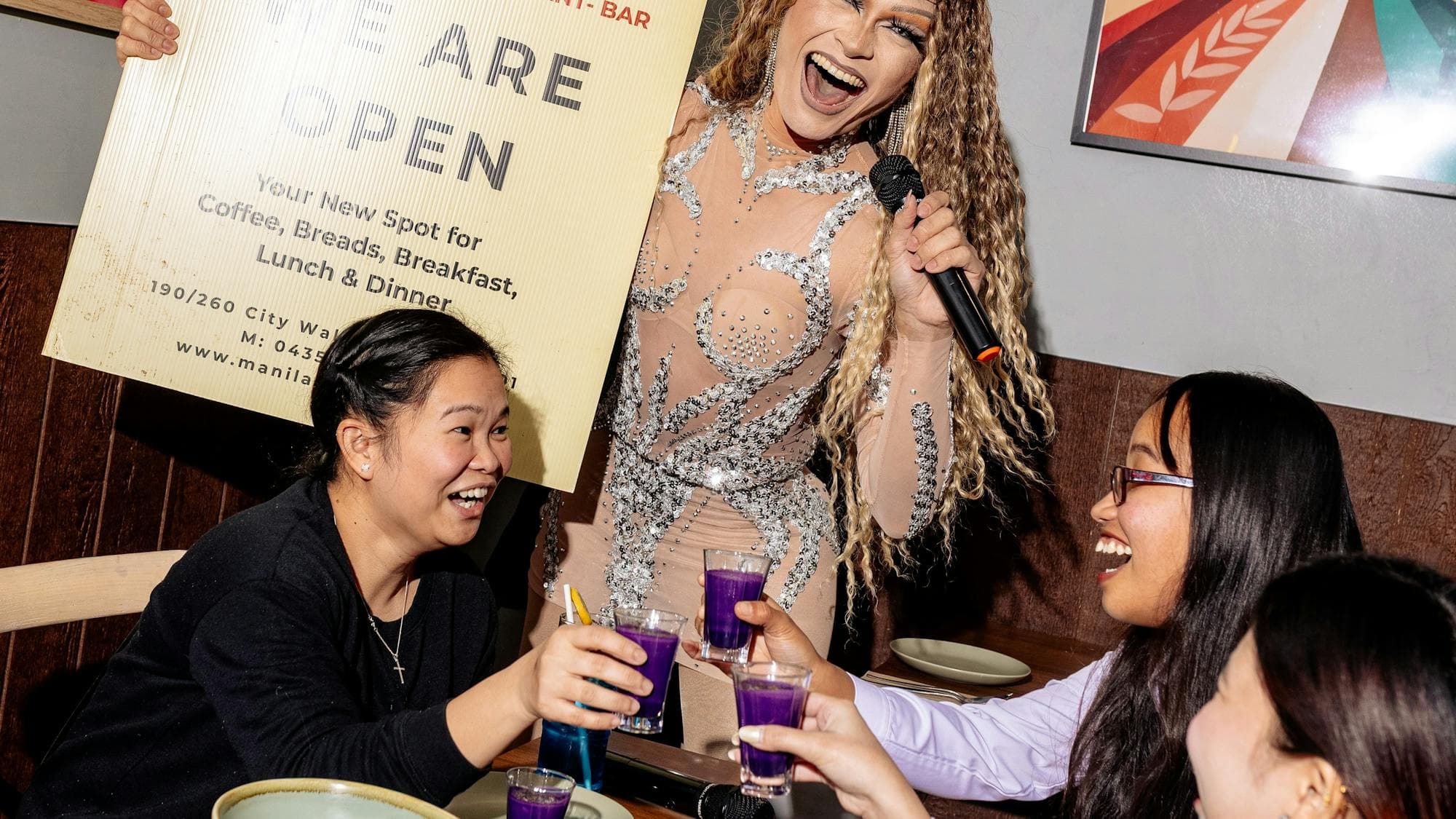 A performer holding a microphone stands beside a table where guests raise purple drinks in a toast,