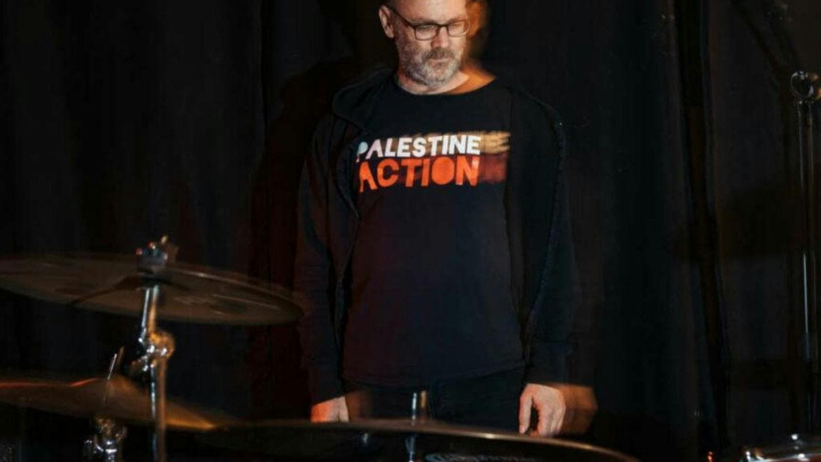 David Rovics standing behind drums with a Palestine Action t-shirt on. Black background.