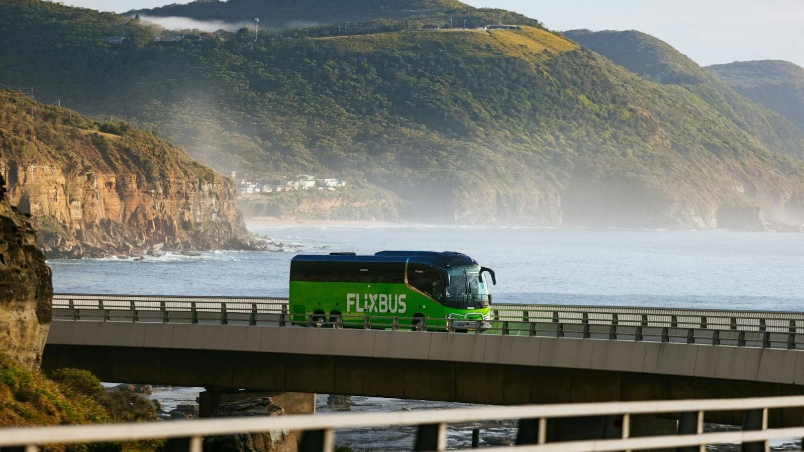 A green flixbus driving on a bridge with sea and mountains in the background.